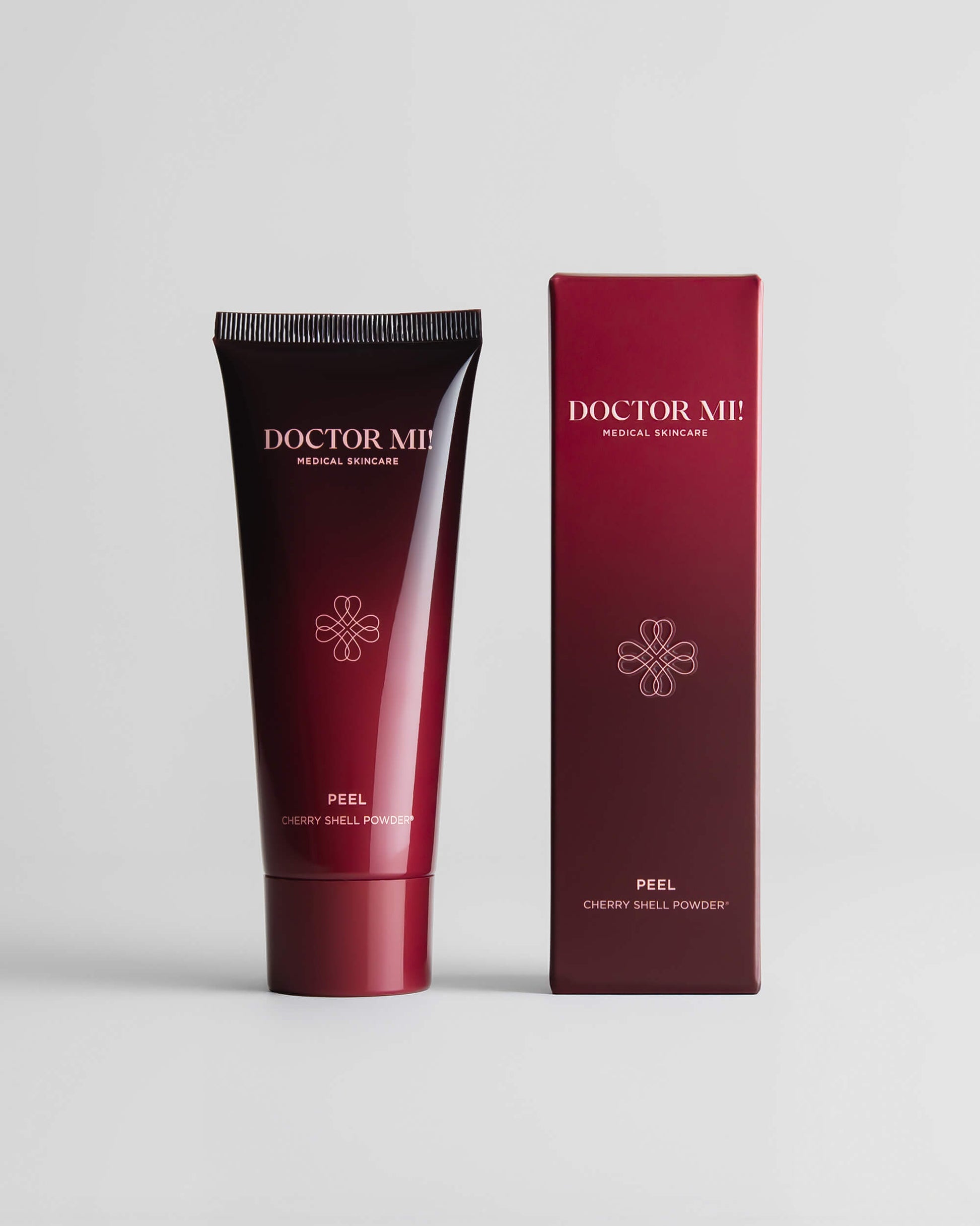 DOCTOR MI! REVIVING PEEL mechanisches und enzymatisches Peeling aus geschroteten Kirschkernschalen in roter Tube neben roter Verpackung auf grauem Hintergrund